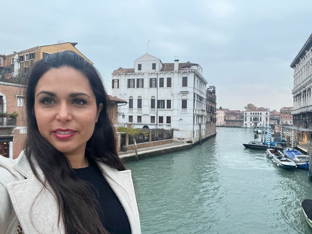 Person posing next to a canal with buildings in the background.
