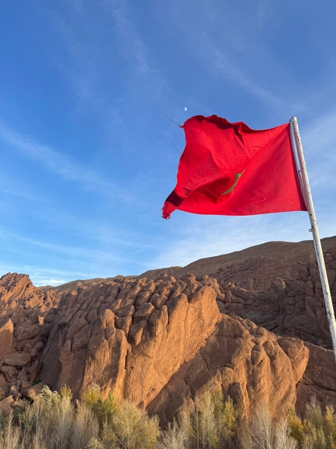       Red Moroccan flag waving against rugged mountain backdrop.
  