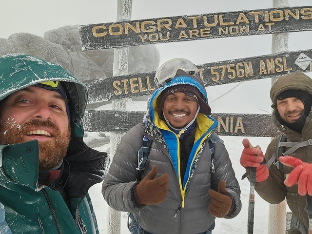       Three people posing at a snowy mountain summit with a sign.
  