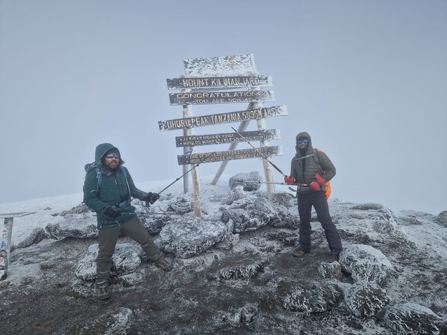       Two people at a snowy mountain summit with signage.
  