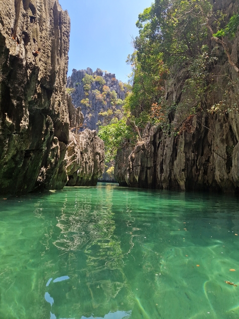       A view of a narrow lagoon flanked by towering rock formations and green foliage.
  