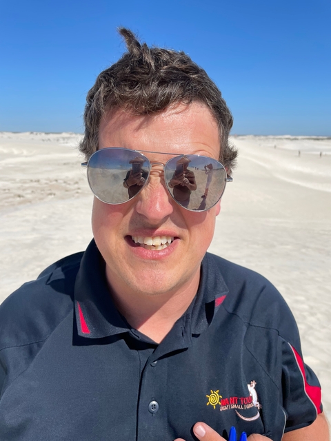 Close-up of a smiling man wearing sunglasses with desert visible in the reflection.
