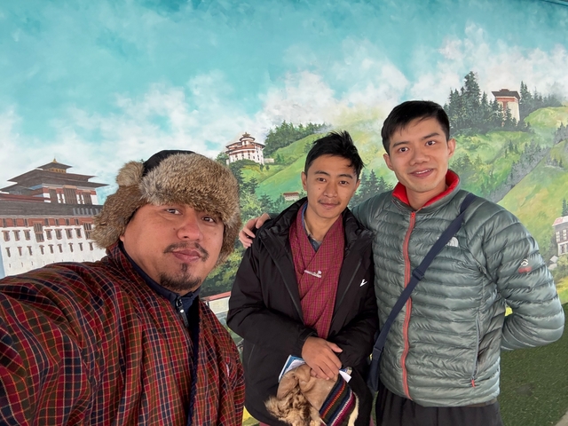       Three smiling men posing in front of a painted backdrop of Bhutanese landscape.
  