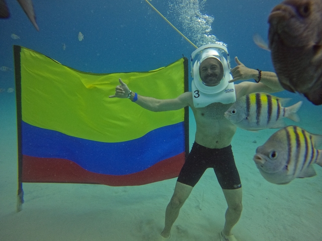 Person underwater with a helmet, a Colombian flag, and fish swimming around.
