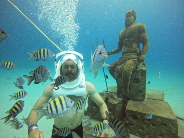 Person underwater with helmet, surrounded by fish and a statue.