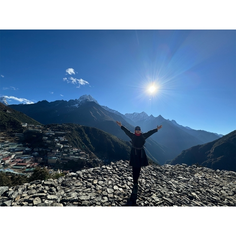       A person standing with arms raised on a hill with mountain vistas.
  