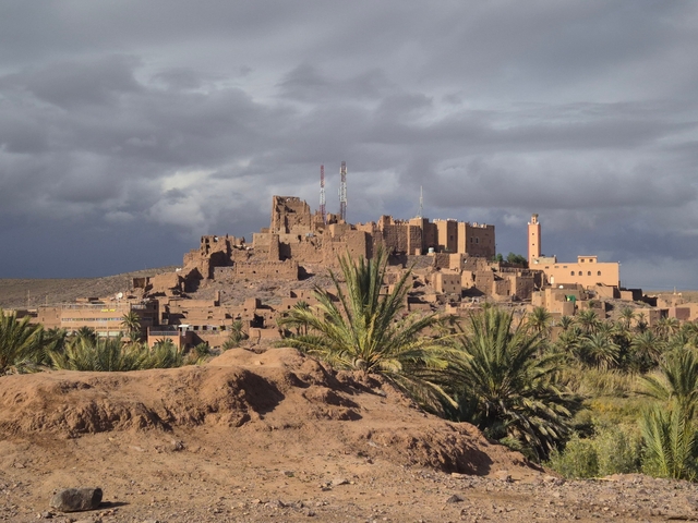       A traditional Moroccan kasbah amidst palm trees under a cloudy sky.
  