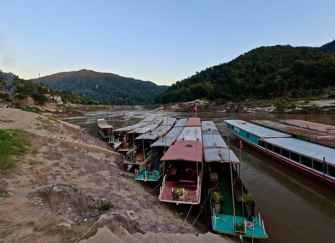 Riverboats docked on a riverbank with forested mountains in the background.