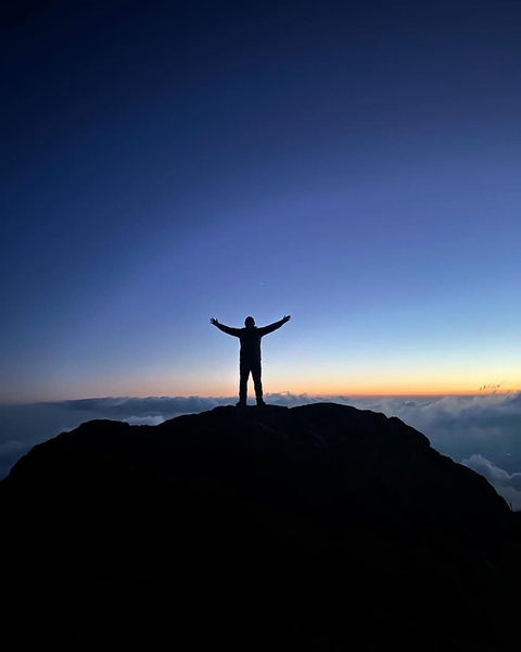 Silhouette of a person on a mountain peak with arms raised at sunrise.