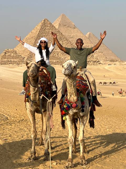       Couple joyfully raising arms on camels with pyramids behind.
  