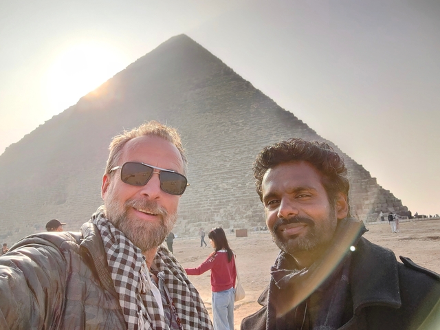 Two men posing in front of a pyramid under the sun.