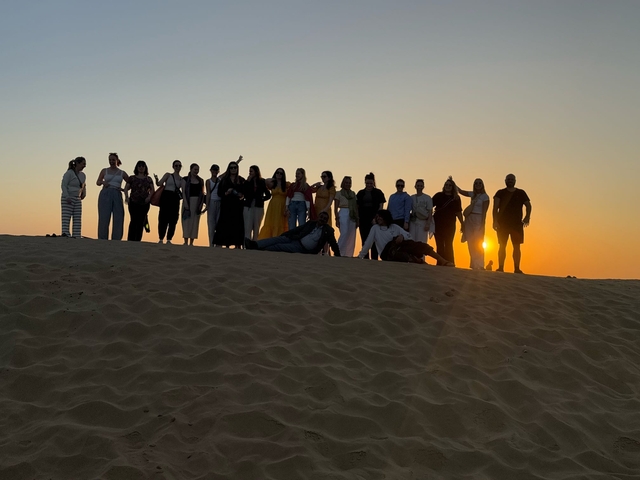       Group of people silhouetted against sunset over sand dunes.
  