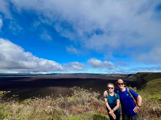       Two people posing in front of a vast landscape with a large crater under a blue sky.
  