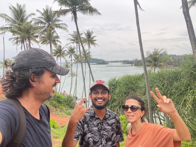 Three people standing on a coastal viewpoint with palm trees and the ocean in the background.