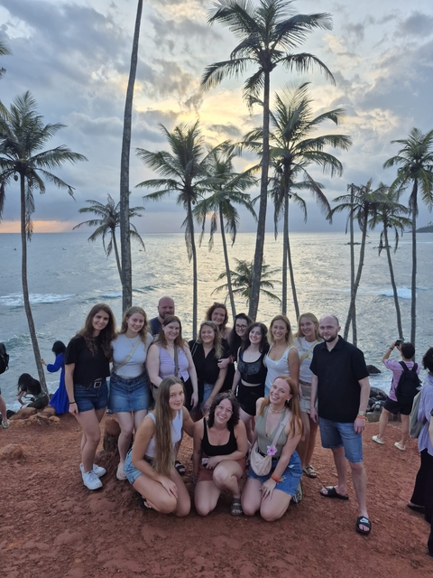 Group of people posing with palm trees and ocean at sunset.