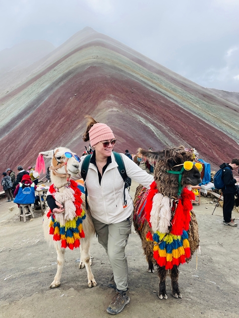       A woman posing with two llamas in colorful attire at Rainbow Mountain.
  