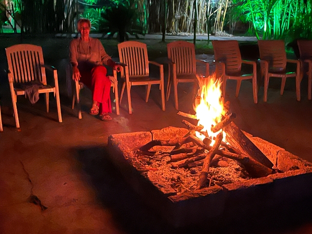       Campfire with empty chairs around and a person sitting close.
  