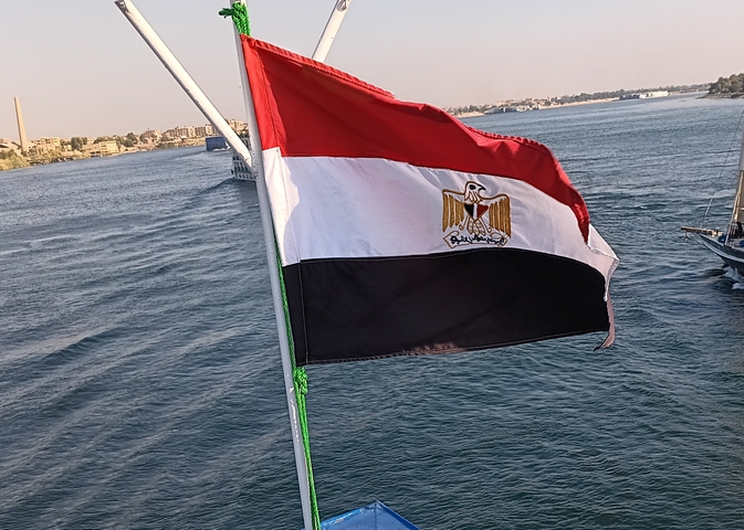       Egyptian flag waving on a ship with a river scene.
  