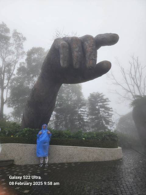 A person stands under a giant stone hand in the mist.