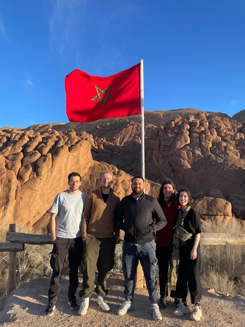       A group of people standing near a Moroccan flag with rocky terrain.
  
