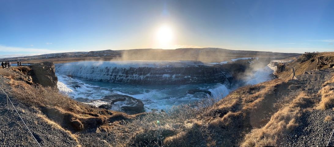       A panoramic view of a waterfall with sunlight and mist.
  