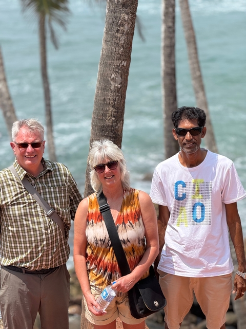       Group of three people standing in front of palm trees and ocean backdrop.
  