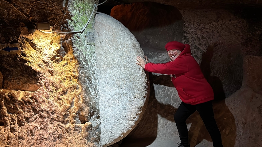       A person in a red outfit posing with a large stone slab in a dim underground setting.
  