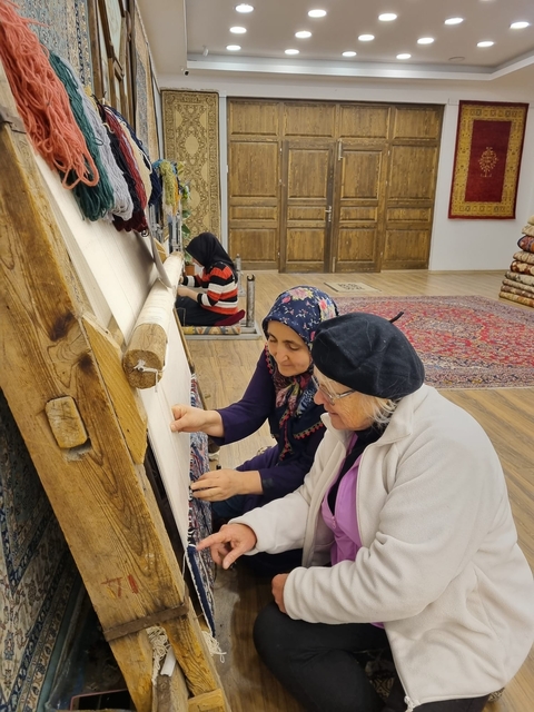       Women weaving a traditional carpet in a cultural setting.
  