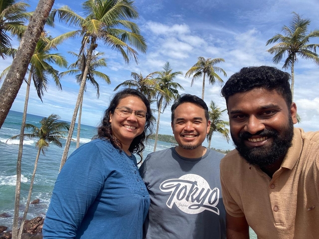 Three people smiling in front of tropical palm trees with ocean view.