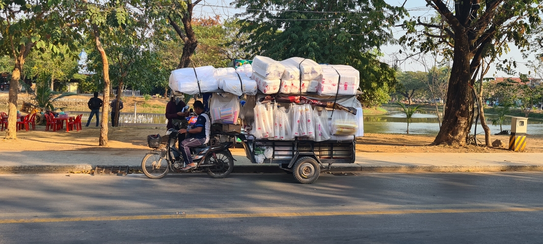       Two people on a motorbike with a heavily loaded trailer.
  