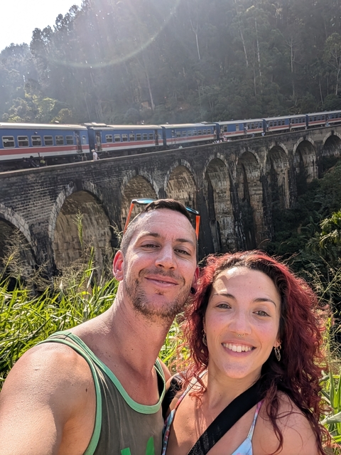       Selfie of a couple with a train on a bridge in the background.
  