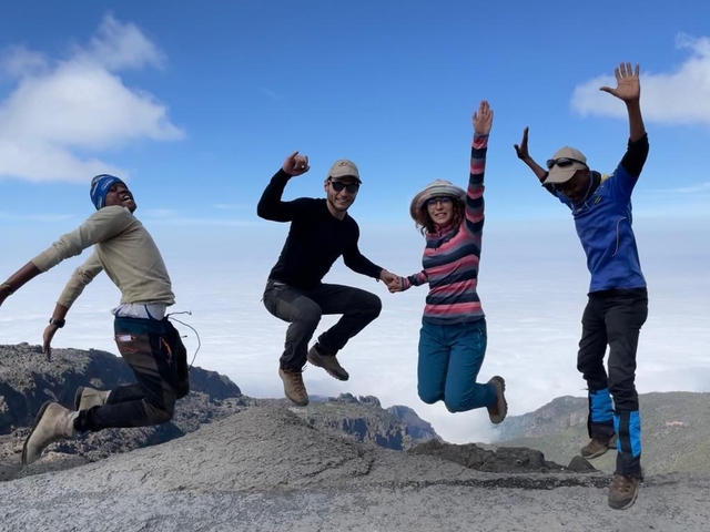 Four people jumping on a mountain with a cloudy sky and distant landscape.