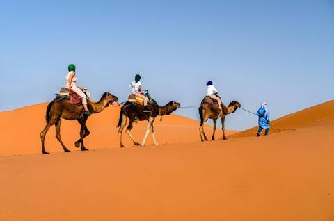 People riding camels through orange desert sands with blue skies.