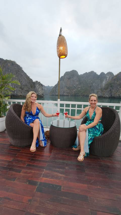 Two women sitting on a deck with drinks, overlooking water and cliffs.