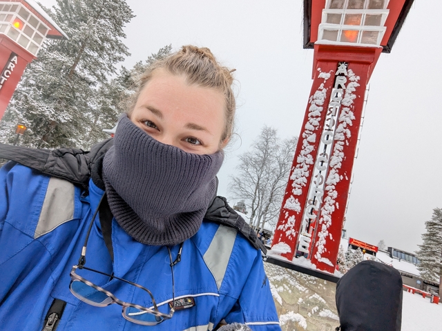       Person in winter clothing near the Arctic Circle sign with snowy trees in the background.
  