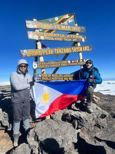Two people holding a flag at Uhuru Peak, Tanzania with a sign in the background.