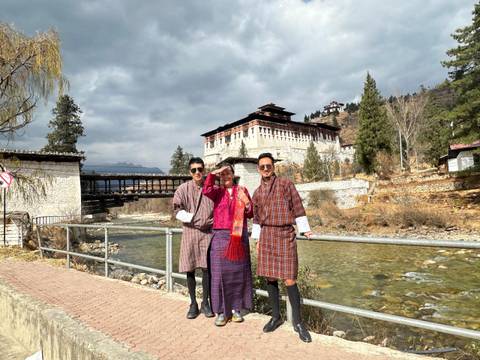 Three people in traditional attire by a river and a bridge.