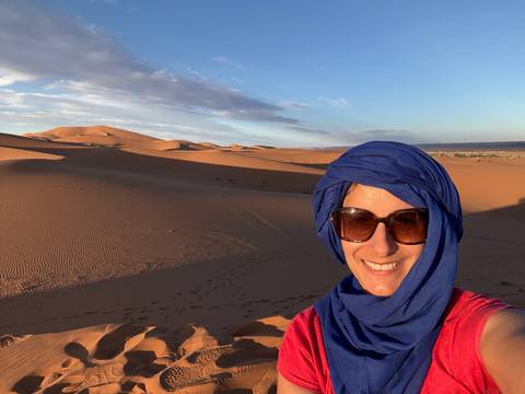       Smiling person in sunglasses and a headscarf with desert sand dunes at sunset in the background.
  
