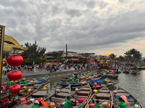       Crowded evening scene by the river with lanterns and boats.
  