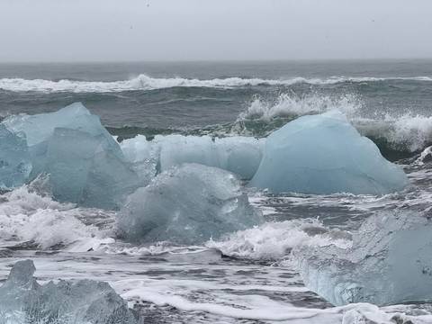 Waves crashing over icebergs on a black sand beach.