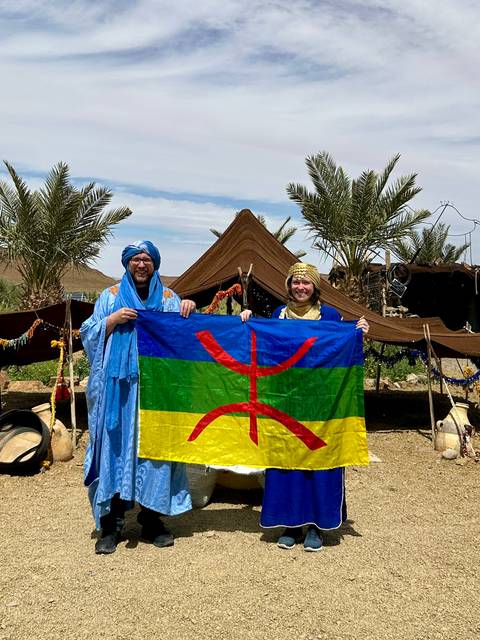       Two people holding a Berber flag in front of a tent.
  