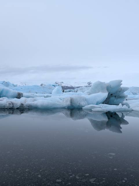       Icebergs floating on calm water under a cloudy sky.
  