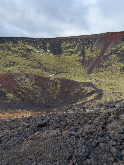       Volcanic crater with red and green hues, under a cloudy sky.
  
