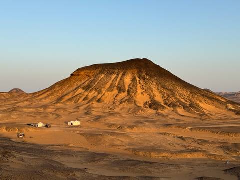       A lone mountain formation in a desert landscape with a few small buildings.
  