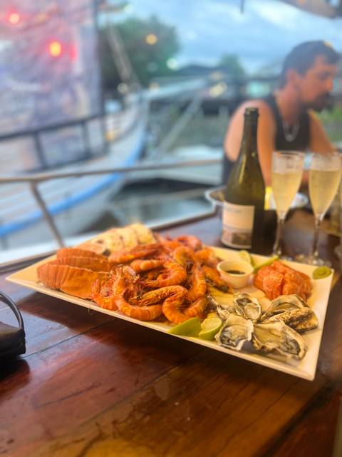       Seafood platter with prawns, oysters, and champagne on a table by the water.
  
