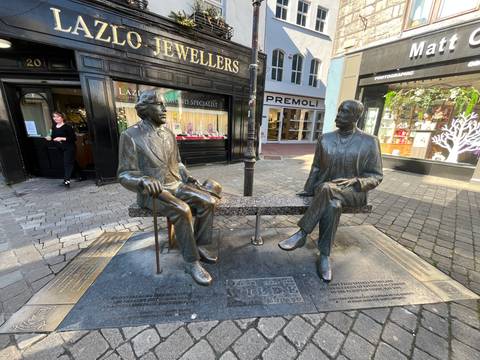       Statues of two seated figures on a street sidewalk.
  