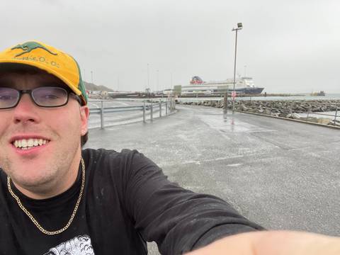 A person taking a selfie at a port with a ferry in the background.