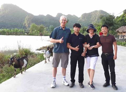 Four people posing with thumbs up in a rural landscape with goats.