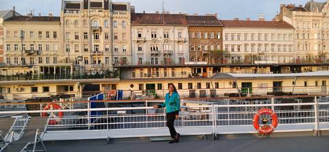 Person posing on a ship with a backdrop of European architecture.