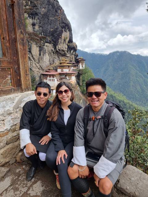       Three people posing with Taktshang Monastery on a cliffside.
  